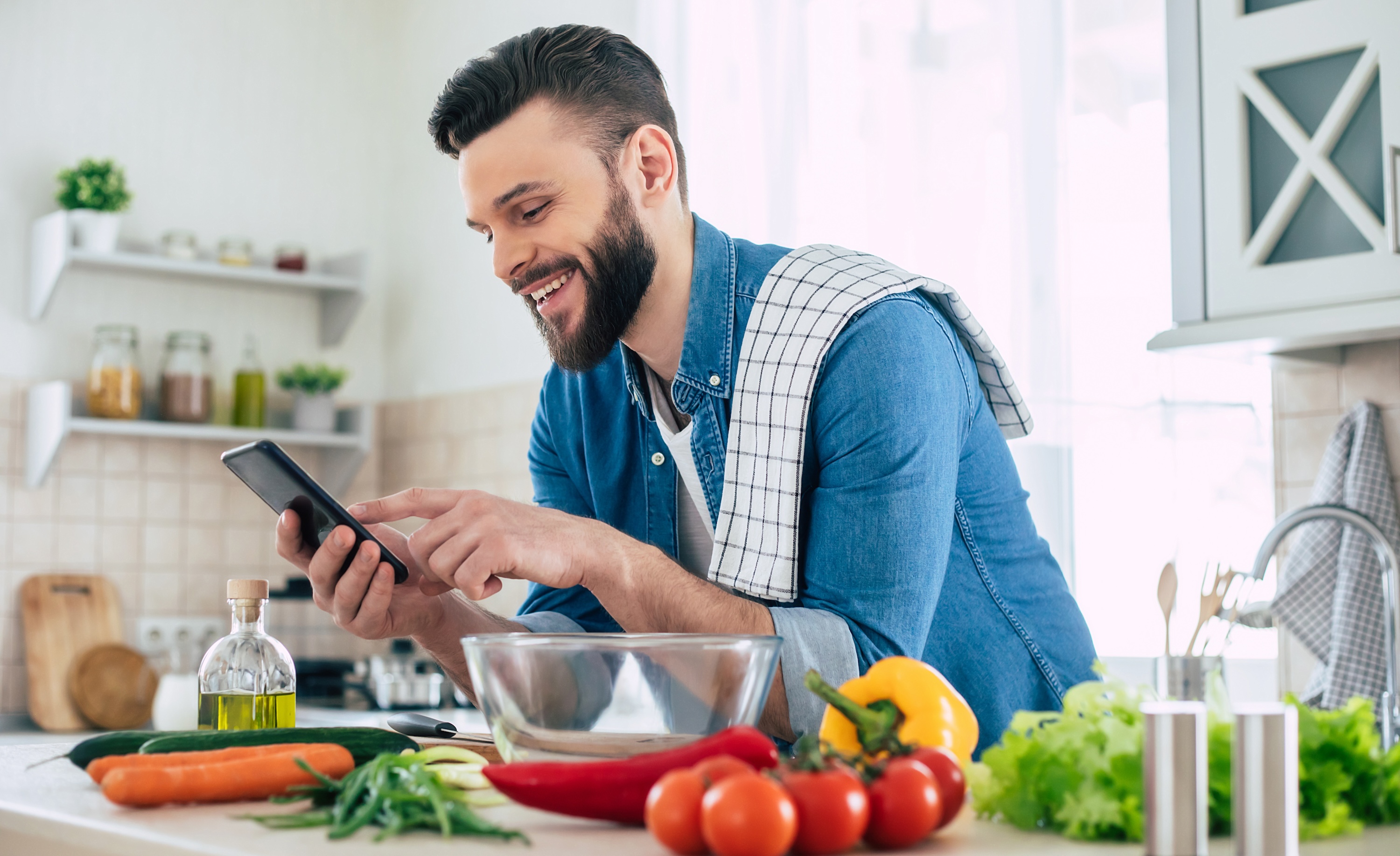 Happy handsome bearded man is using his smart phone while he preparing vegan healthy breakfast for a lovely family